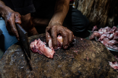 A butcher is cutting a freshly slaughtered chicken into bite size pieces for the customer. An old, thick wooden stump is used as the table for the cutting. India, 2021. S. Chakrabarti / We Animals