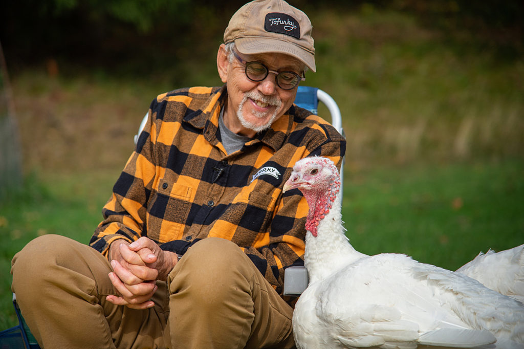 Tofurky founder Seth Tibbott and rescued turkeys enjoy a meal and each other's company at Wildwood Farm Sanctuary & Preserve. USA, 2021. Jo-Anne McArthur / We Animals