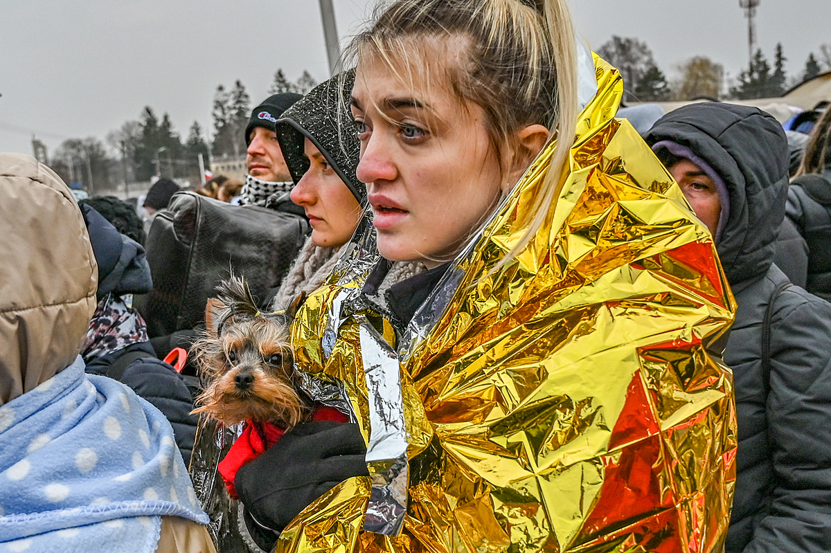 A Ukrainian refugee holds her small dog tightly against her chest as she waits in line at the reception center in Medyka on the Polish Ukrainian Border. An emergency blanket is wrapped around her shoulders to help her stay warm from the bitter winter cold. Poland, 2022. Miloš Bičanski / We Animals